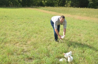 Éleveur Le Gaulois souriant reposant une volaille sur l'herbe de son exploitation et entouré de trois autres poulets