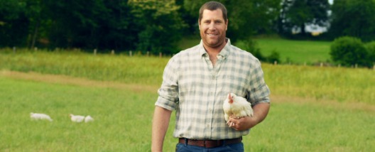 Éleveur Le Gaulois souriant tenant un poulet dans ses bras dans son champs à l'extérieur du poulailler