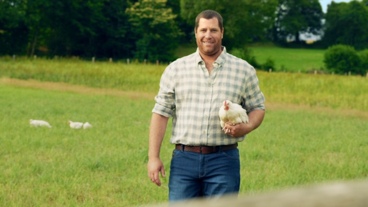 Éleveur Le Gaulois souriant tenant un poulet dans ses bras dans son champs à l'extérieur du poulailler